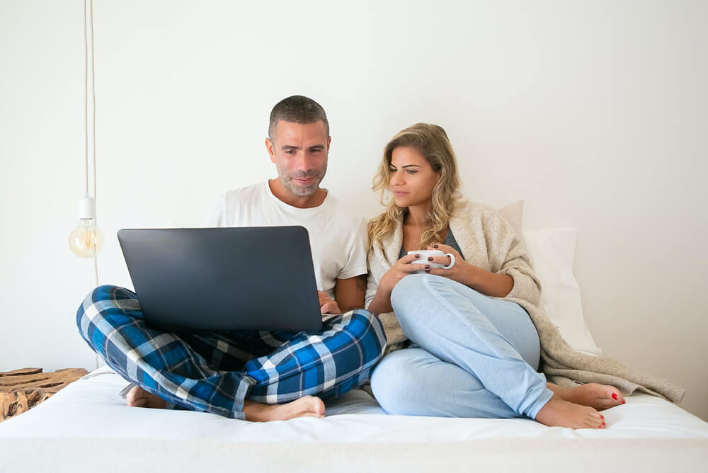 white couple sitting on bed looking at laptop white couple sitting on bed looking at laptop